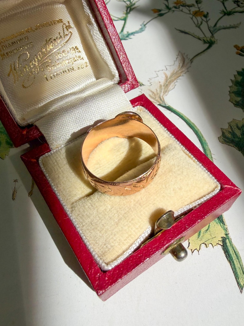 May include: A gold ring with an engraved floral pattern on the outer band, resting inside a red rectangular jewelry box with a cream-colored velvet interior. The box lid features gold embossed text that reads "Viney & Sons Ltd".
