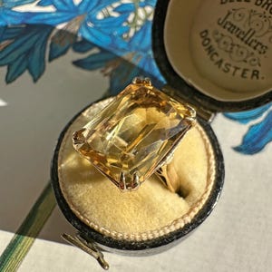 May include: A gold ring with a large, rectangular, yellow gemstone, displayed in a black jewelry box with the text "BELL BROS Jewellers DONCASTER". The ring is set against a yellow velvet interior.