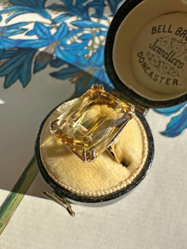 May include: A gold ring with a large, rectangular, yellow gemstone, displayed in a black jewelry box with the text "BELL BROS Jewellers DONCASTER". The ring is set against a yellow velvet interior.