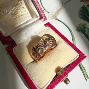 May include: A gold ring shaped like a belt buckle with an engraved floral pattern on the band. The ring is displayed inside a red, rectangular, hinged jewelry box with a cream-colored velvet interior lining and gold-colored text on the lid.
