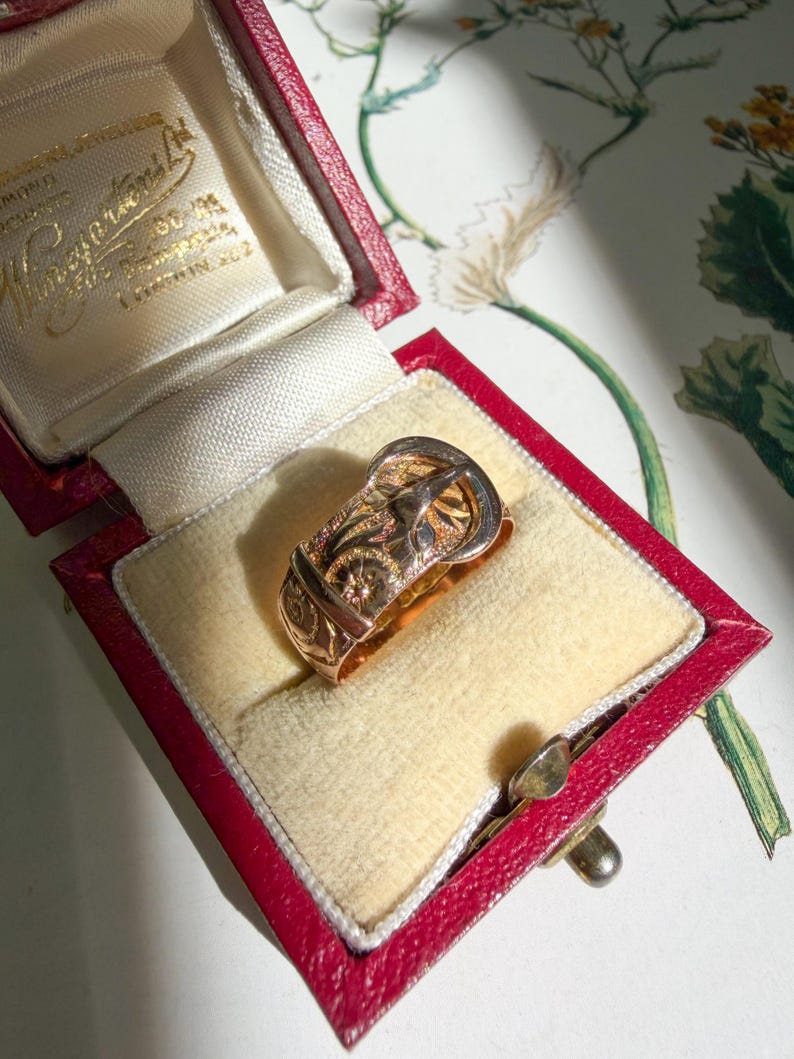 May include: A gold ring shaped like a belt buckle with an engraved floral pattern on the band. The ring is displayed inside a red, rectangular, hinged jewelry box with a cream-colored velvet interior lining and gold-colored text on the lid.