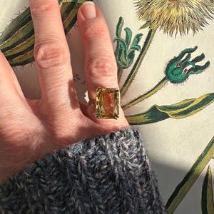 May include: A gold ring with a large, rectangular, light-yellow gemstone. The ring is worn on a finger, set against a botanical illustration with green and yellow hues. The person is wearing a blue and gray knit sweater.