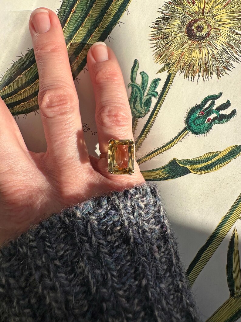 May include: A gold ring with a large, rectangular, light-yellow gemstone. The ring is worn on a finger, set against a botanical illustration with green and yellow hues. The person is wearing a blue and gray knit sweater.