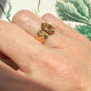 May include: A gold-toned ring featuring a buckle-style design with an engraved leaf pattern along the band. The metal surface displays a polished finish with intricate detailing on the buckle component and the textured band surrounding the finger.
