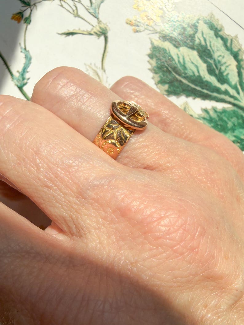 May include: A gold-toned ring featuring a buckle-style design with an engraved leaf pattern along the band. The metal surface displays a polished finish with intricate detailing on the buckle component and the textured band surrounding the finger.