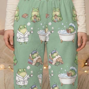 May include: Light green pajama trousers featuring a repeating pattern of cartoon frogs in relaxing poses. Some frogs wear bathrobes, others are in bathtubs, and some read books. The trousers have a comfortable, loose fit.