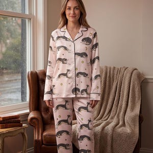 May include: A light pink pajama set featuring a repeating pattern of grey and white dogs in various resting positions, accented with small gold stars and moons. The long-sleeved button-down top and matching trousers have dark piping along the edges.