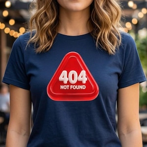 Pode incluir: Camiseta azul marinho com um gráfico vermelho e branco. O gráfico é um triângulo vermelho com o número "404" e as palavras "NOT FOUND" em branco. A camiseta é combinada com jeans pretos.
