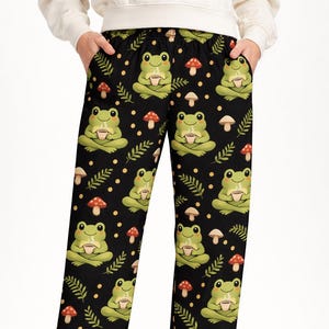 May include: Black pajama trousers featuring a repeating pattern of green frogs holding cups, red and white mushrooms, and sprigs of leaves. The frogs are in a seated position. The background is black with small yellow dots.