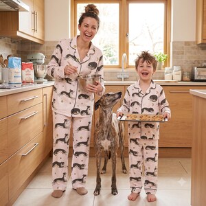 May include: Matching light pink pajama sets featuring a repeating pattern of black and tan greyhounds, small stars, and moons. The pajamas consist of long-sleeved button-down shirts with collars and matching long pants with dark piping along the edges.