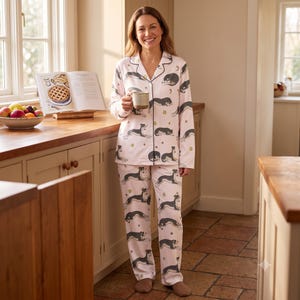 May include: A light pink pajama set featuring a repeating pattern of black and white dogs in various resting positions, accented with small stars and moons. The long-sleeved button-down top has a notched collar and dark piping along the edges.