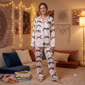 May include: A light pink pajama set featuring a repeating pattern of black and white greyhounds in various resting positions, accented with small gold stars and circles. The long-sleeved button-down top has a notched collar and black piping trim.