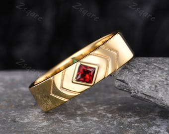 May include: A gold-colored ring with a square, red gemstone set in the center. The ring has a geometric design with chevron patterns. The ring is displayed on a gray, textured surface.