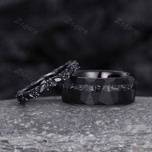 Könnte beinhalten: Ein Paar schwarze Eheringe. Ein Ring ist ein schlichtes, breites Band mit einer strukturierten Oberfläche. Der andere Ring ist ein dünnes Band mit schwarzen Edelsteinen.