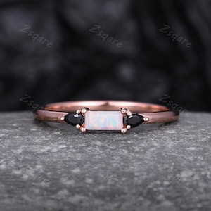 May include: Rose gold ring featuring a rectangular opal center stone flanked by two black onyx stones. The ring has a delicate band and is set on a gray surface. The ring is a piece of jewelry.