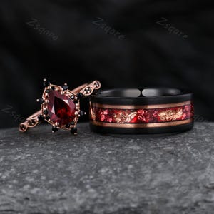 May include: A rose gold engagement ring featuring a pear-cut red garnet stone surrounded by small black stones, paired with a black band featuring a central inlay of crushed red and gold-toned material. The text Zsqare is visible in the background.