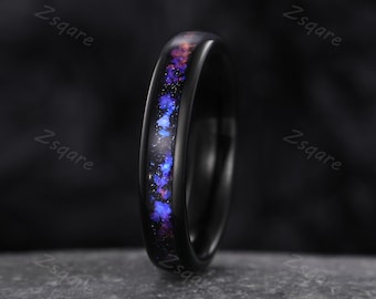 May include: A black ring featuring a colourful inlay of blue, purple, and orange, reminiscent of a galaxy. The ring's band is solid black, providing a strong contrast with the vibrant interior. The ring is set against a dark backdrop.