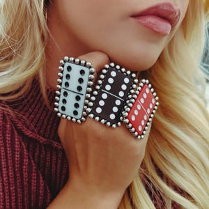 May include: Three large domino rings in a hand. The rings are white, black, and red, each with a different dot pattern. The rings are framed with small silver beads. The person is wearing a burgundy sweater.