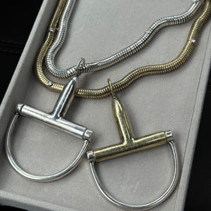 May include: Two necklaces with horse bit pendants. One pendant is silver, the other is gold. The necklaces have a snake chain design, with one silver and one gold. The jewelry is displayed in a grey box.