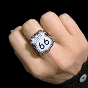 May include: A Route 66 sign-shaped ring with a white background and black text. The ring features the words "Route 66" and the number "66". The ring is surrounded by a border of small, dark-colored stones.