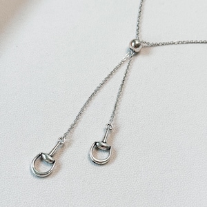 May include: A silver-toned lariat necklace featuring a delicate cable chain and a sliding bead mechanism. Two dangling ends are each finished with a small, metallic horse bit charm, creating a symmetrical drop design with a polished finish.