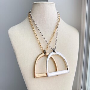 May include: Two necklaces featuring stirrup-shaped pendants hanging from matching link chains. One pendant and chain are finished in a polished gold tone, while the other pendant and chain are finished in a polished silver tone. The pendants are smooth.