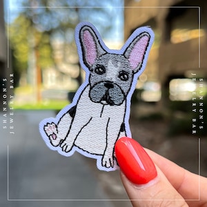 Op de afbeelding: Een geborduurde patch van een zittende Franse bulldog met zichtbare poten. De patch is wit en grijs en bevindt zich op een witte achtergrond.