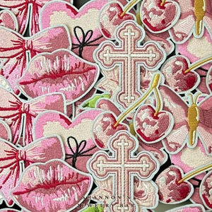 May include: A collection of pink and red embroidered iron-on patches featuring various designs, including lips, bows, cherries, crosses, and butterflies. The patches are all in a pink and red colour scheme and have a vintage aesthetic. The text "SHANNON'S JEWELRY BAR" is visible at the bottom of the image.