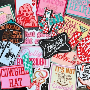 May include: A collection of colourful iron-on patches with various designs, including a cowboy hat, a lightning bolt, a heart, a horseshoe, and the words "Howdy Cowboy", "Cowgirl Hat", "Talk Dirty To Me", "Hot Girl Summer", "It's Not Easy Being A Cowgirl", "Dolly For President", and "Do No Harm But Take No Bull".