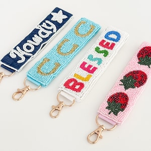 May include: Four beaded keychains with gold-coloured clasps. One is denim blue with "Howdy" in white beads, another is light blue with gold horseshoe beads, a third is white with "Blessed" in colourful beads, and the last is pink with red strawberry beads.