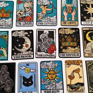 Puede incluir: Una colección de parches bordados de cartas del tarot. Los parches presentan varios diseños, incluyendo esqueletos, coronas, lunas y soles, con textos como "The Lovers" y "The Moon". La paleta de colores incluye negro, azul, dorado y rojo.