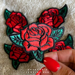 May include: Embroidered red rose patches with green leaves and black outlines. The patches feature detailed stitching and are suitable for fabric embellishment. The image also includes the text "SHANNON'S JEWELRY BAR".