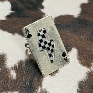 Pode incluir: Anel prateado com um design de ás de espadas. O anel apresenta uma carta de baralho com um raio em padrão xadrez preto e branco. O anel está sobre um padrão de pele de vaca.