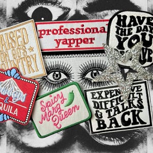 May include: An assortment of embroidered patches with text and designs. The patches feature phrases such as "professional yapper" and "Spicy Marg Queen." A silver star-shaped embellishment is also visible. The background has a black and white eye design.