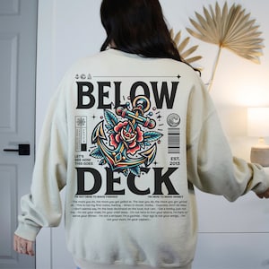 Op de afbeelding: Een beige sweatshirt met een grafische print van een anker met een rode roos en de tekst "BELOW DECK" in zwart. De tekst "LET'S SEE HOW THIS GOES" en "EST. 2013" zijn ook op het sweatshirt gedrukt.
