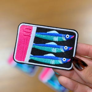May include: Embroidered patch featuring three stylized sardines in a can. The sardines are blue and teal with white accents and pink mouths, set against a black background. The can is pink with white and black trim.