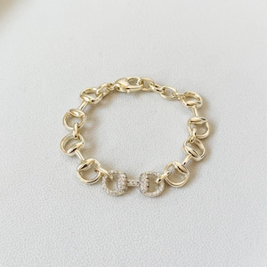 May include: A gold-toned chain link bracelet featuring a series of repeating equestrian-inspired bit motifs. The central link is embellished with small, clear pavé-set stones, while the remaining links have a smooth, polished metallic finish.
