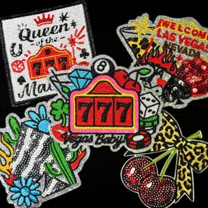 Puede incluir: Un conjunto de parches bordados con temática de Las Vegas. Los diseños incluyen una máquina tragamonedas con el número 777, un letrero de bienvenida, dados, cerezas y un lazo con estampado de leopardo. Los parches están adornados con lentejuelas.