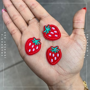 May include: Three red embroidered strawberry patches with green leaves and white seeds. The patches are on a hand.