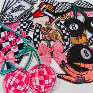 May include: A collection of colorful embroidered iron-on patches featuring various designs, including cherries, horseshoes, a cowboy girl, a 8-ball, and a hand with a cigarette. The patches are perfect for adding a unique touch to clothing, bags, or other accessories.  The text "Shannon's Jewelry Bar" is visible at the bottom of the image.