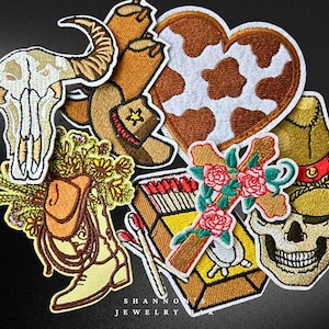 May include: A set of 10 embroidered iron-on patches featuring various western and cowboy themes. The patches include a skull, a cowboy hat, a heart with a cowhide pattern, a cross with roses, a boot with a sunflower bouquet, a box of matches, and a skull with a cowboy hat.