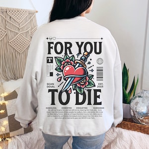 Könnte beinhalten: Weißer Pullover mit einem grafischen Druck eines Herzens, das von einem Dolch mit Blumen durchbohrt wird. Der Text "For You To Die" ist in schwarzen Buchstaben gedruckt. Der Text "Demented, Disgusting, Subhuman, Diabolical" ist in kleineren Buchstaben unterhalb der Grafik gedruckt. Der Text "Est. 2023" ist in der rechten unteren Ecke gedruckt.