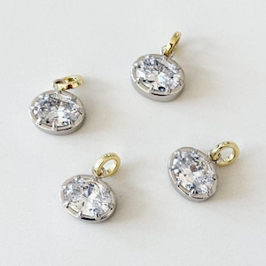 May include: Four oval-shaped pendants with a silver-colored setting and a gold-colored bail. Each pendant features a large, clear, faceted stone, creating a sparkling effect. The pendants are displayed on a white surface.