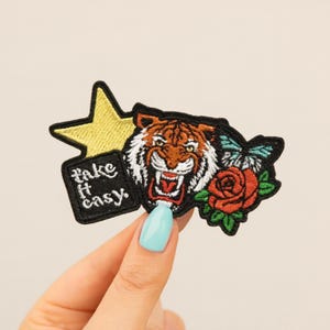May include: Embroidered patch featuring a roaring tiger, a red rose, and a butterfly. The patch includes a yellow star and a black rectangle with the words "take it easy." The patch is black with colorful accents.