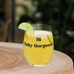 Può includere: Un bicchiere di vino in vetro trasparente con una bevanda gialla e una fetta di lime. Il bicchiere ha la scritta "Hi Baby Gorgeous" stampata su di esso.