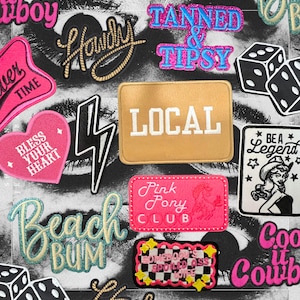 May include: An assortment of embroidered patches with diverse designs and text. Patches include phrases such as "Howdy," "Tanned & Tipsy," and "Beach Bum." Other patches feature a lightning bolt, dice, and a pink heart with "Bless Your Heart."