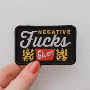 May include: Black rectangular embroidered patch with the words "NEGATIVE FUCKS GIVEN" in white and yellow lettering. The patch has a red rectangle with the word "Given" in white. Two yellow decorative emblems are on either side of the red rectangle.