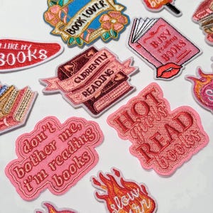 以下が含まれることがあります： 本をテーマにしたデザインの刺繍パッチのコレクション。パッチには「Book Lover」、「Currently Reading」、「Hot Reads Books」などのフレーズが使用されています。カラーパレットはピンク、赤、青で、花や本のイラストが描かれています。