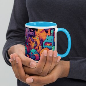 May include: A white ceramic mug with a blue rim and handle. The mug features a colourful illustration of cartoon monsters with a dark background.