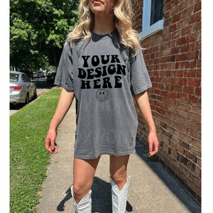 May include: A gray oversized t-shirt with the text "YOUR DESIGN HERE" and a smiley face graphic. The model is wearing white cowboy boots.